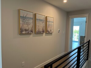 A hallway with three framed artworks on the wall, a thermostat, and a view into an adjacent room. - Life is Better at the Beach! (Wildwood)