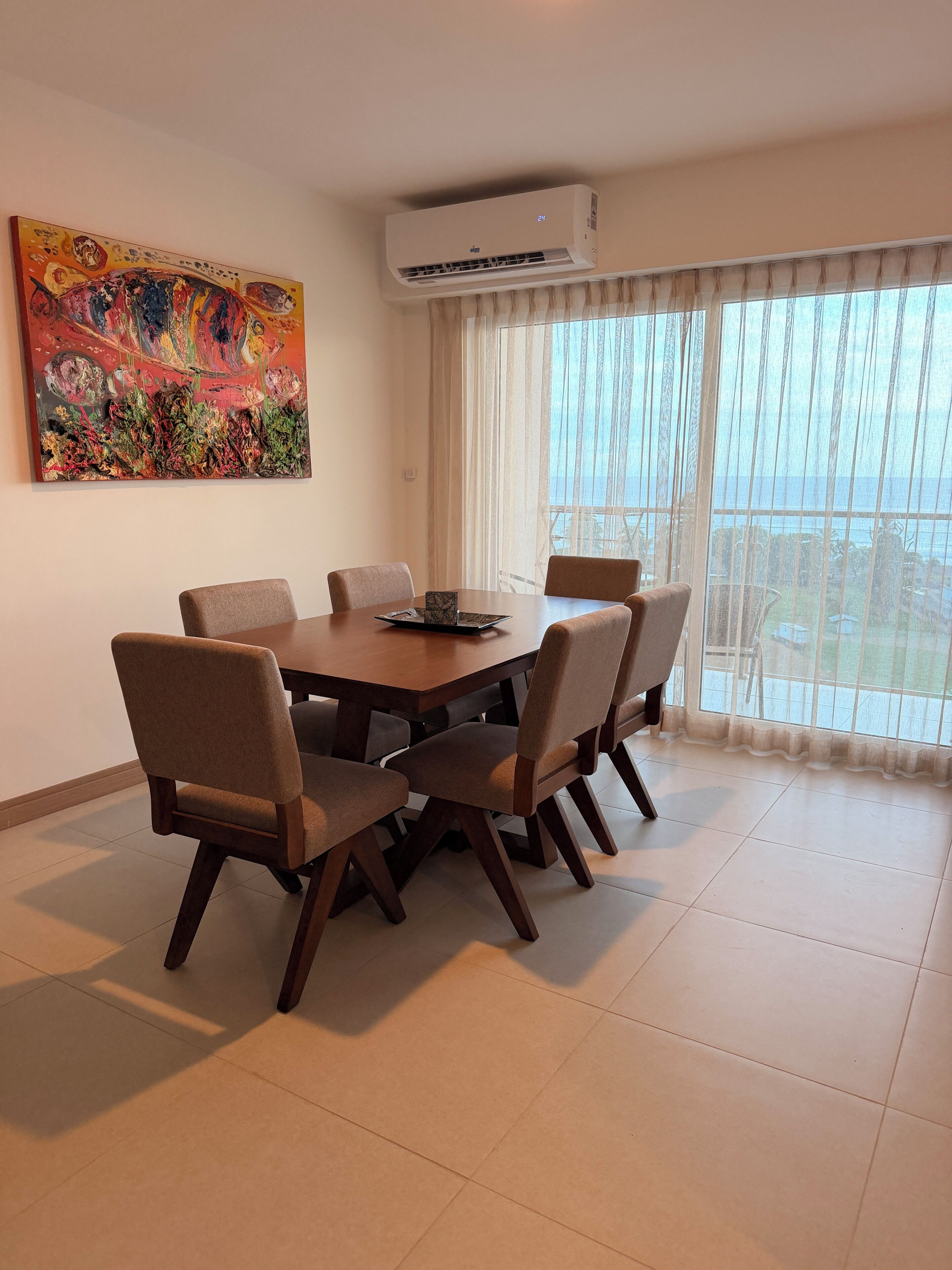 Uma sala de jantar com mesa redonda, seis cadeiras, ar condicionado, um grande quadro abstrato e vista através de cortinas leves.