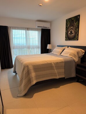 Une chambre à coucher comprenant un lit, une table de chevet, une lampe, une fenêtre avec des rideaux et un appareil de climatisation.