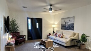 A living room with a sofa, a coffee table, a TV, a ceiling fan, a door, and a painting on the wall. - Spacious 2BR Guest House - Perfect for Remote Work, Family and Pet-Friendly (Las Vegas)