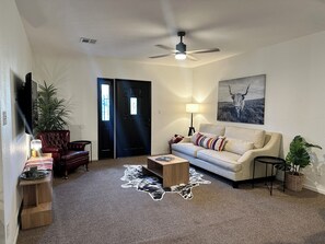 A living room with a sofa, a coffee table, a TV, a ceiling fan, a door, and a painting on the wall. - Spacious 2BR Guest House - Perfect for Remote Work, Family and Pet-Friendly (Las Vegas)