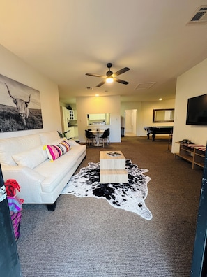 A living room with a white sofa, a cowhide rug, a wooden coffee table, a ceiling fan, a kitchen in the background, and a TV mounted on the wall. - Spacious 2BR Guest House - Perfect for Remote Work, Family and Pet-Friendly (Las Vegas)