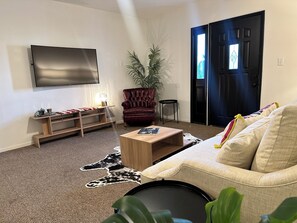 A living room with a flat-screen TV, a leather armchair, a sofa, a coffee table, a plant, and a door with a window. - Spacious 2BR Guest House - Perfect for Remote Work, Family and Pet-Friendly (Las Vegas)
