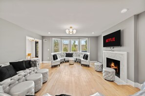 Una sala de estar moderna con un sofá seccional, otomanas, una chimenea y un televisor mostrando 'NETFLIX'.
