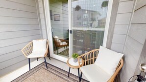 A balcony with two wicker chairs and a small table, a sliding glass door, and a view of a building. - *New* Ducks & Diamonds 3br/3bath (Bryan)