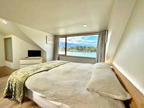 Ein Schlafzimmer mit einem großen Bett, einem Fernseher auf einem Schrank und Blick auf einen See und Berge.