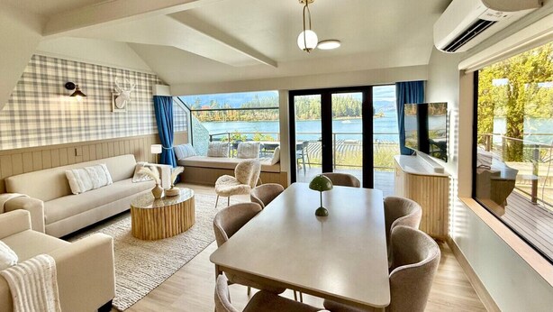 Een moderne woonkamer met een bank, salontafel en eettafel. Er is een groot raam met uitzicht op een meer.