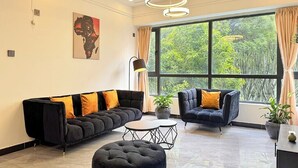 Ett modernt vardagsrum med en svart soffa, en svart fåtölj, en rund pall och ett litet bord. Det finns ett stort fönster med gardiner, en tavla på väggen och en taklampa.