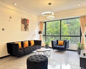 Ein modernes Wohnzimmer mit einem schwarzen Sofa, einem schwarzen Sessel, einem runden Hocker und einem kleinen Tisch. Es gibt ein großes Fenster mit Vorhängen, ein Bild an der Wand und eine Deckenleuchte.