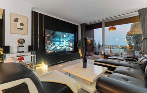 A modern living room with a large flat-screen TV, leather sofas, a coffee table, and a view of the city through large windows.