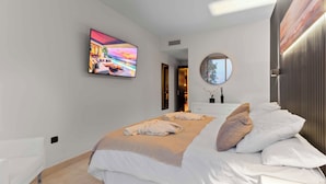 A modern hotel room with a large bed, a flat-screen TV, a round mirror, and a view of the sea.