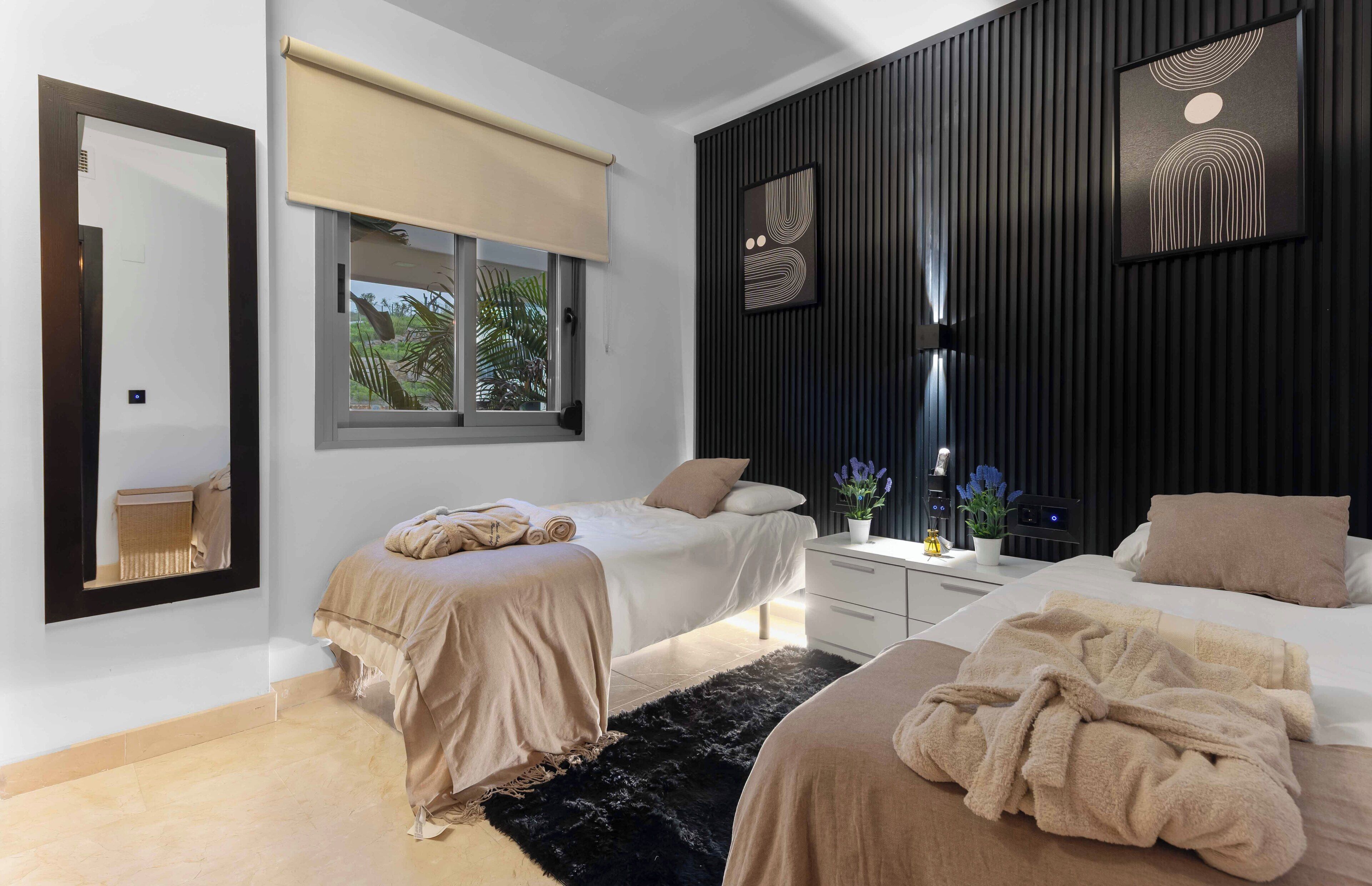 Un dormitorio moderno con dos camas, un espejo grande, una ventana con persiana, una pared negra con textura y una mesita de noche con una planta.