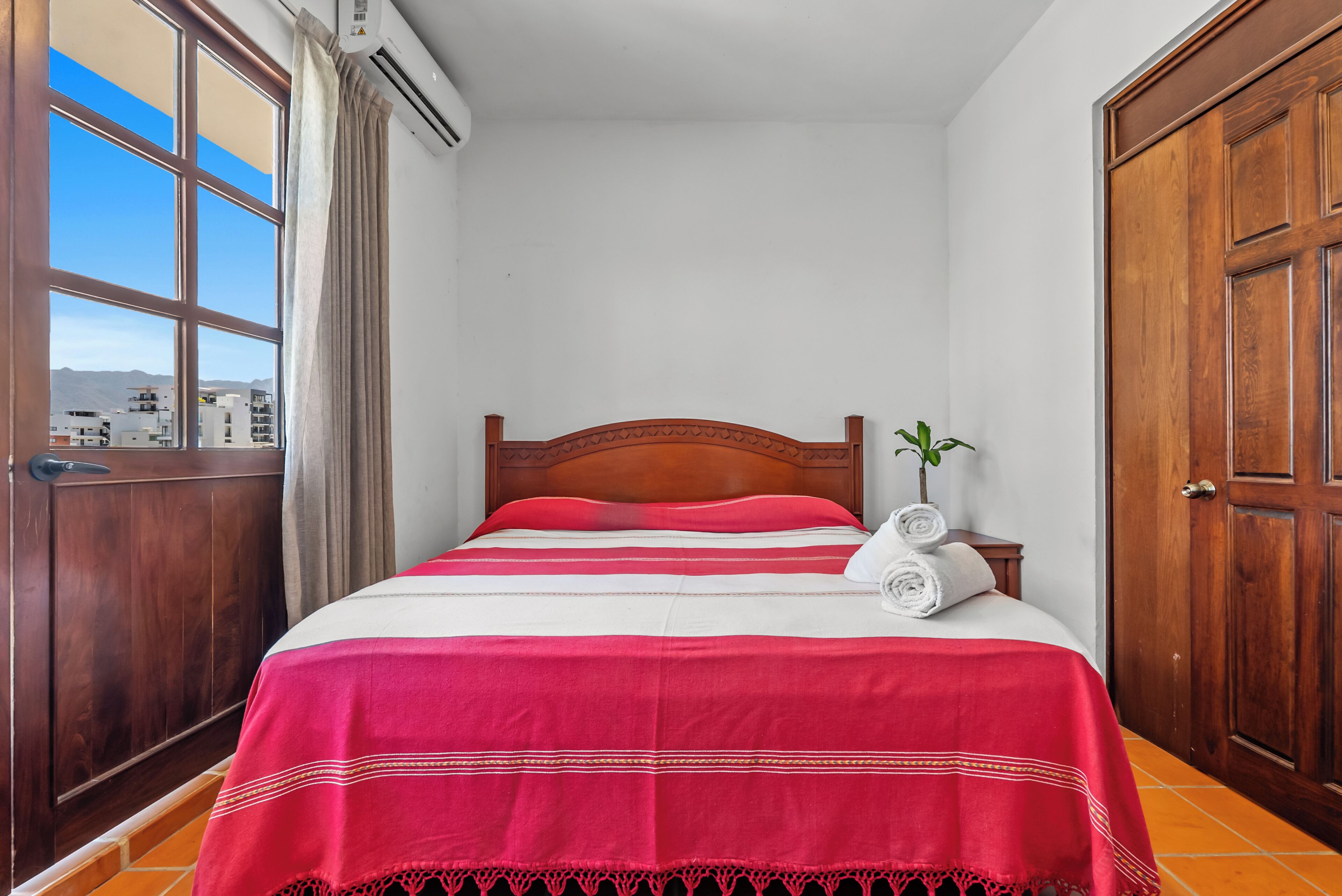 A neatly made bed with a red and white bedspread, a wooden headboard, and a view of buildings through a window with curtains.