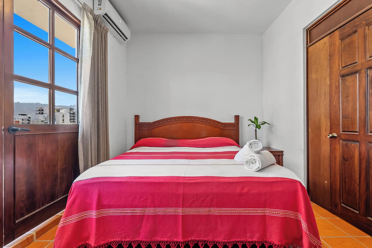 Una cama bien tendida con colcha roja y blanca, cabecera de madera y vistas a edificios a través de una ventana con cortinas.