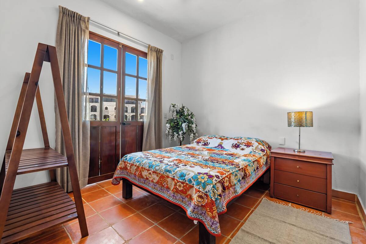 A bedroom with a bed, a wooden ladder, a nightstand, a lamp, and a view of a building through a window.