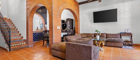 A living room with a brick archway, a brown sofa, a TV mounted on the wall, and a staircase with patterned tiles.