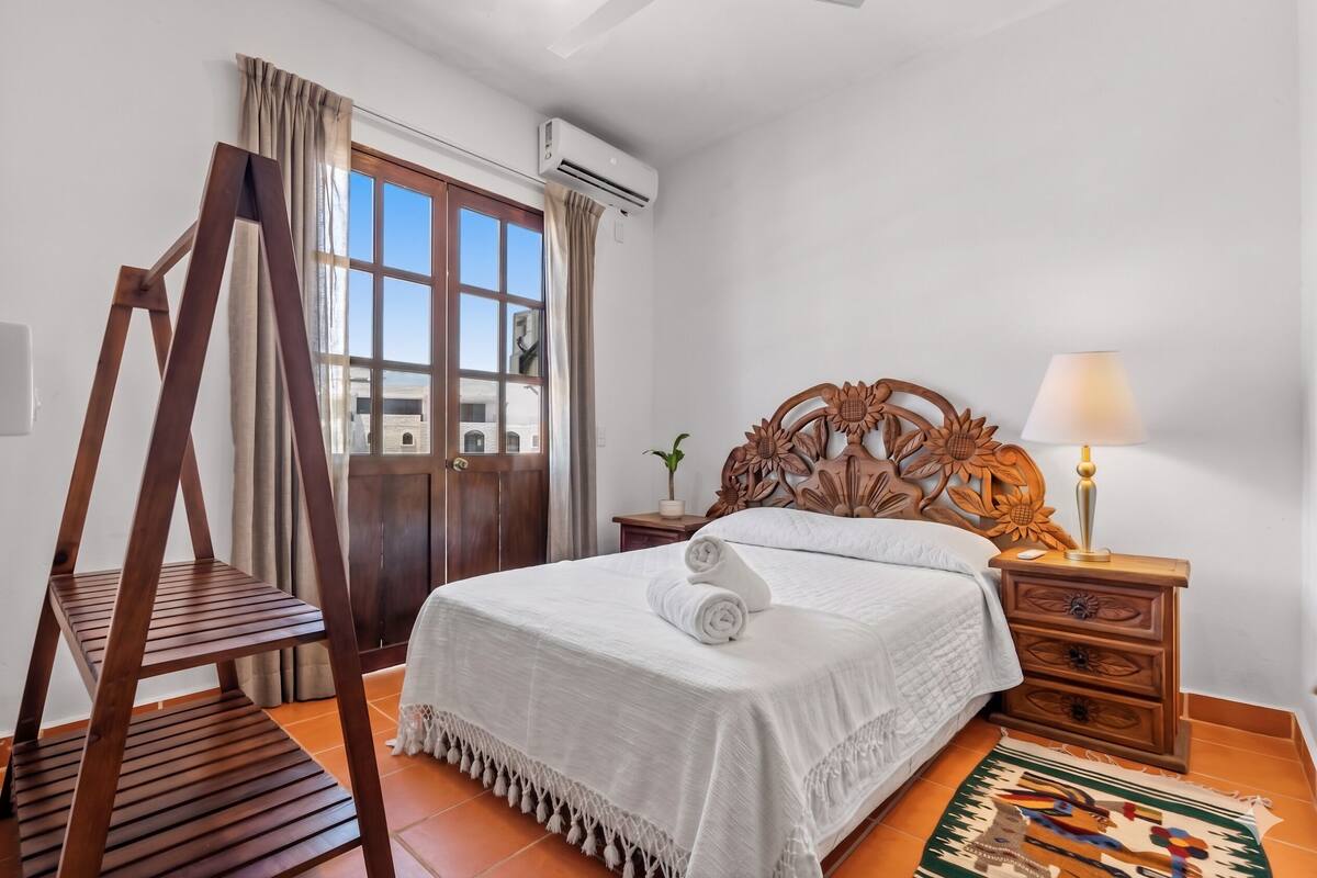 A bedroom with a bed, wooden nightstand, lamp, air conditioning unit, window with curtains, and a wooden ladder.