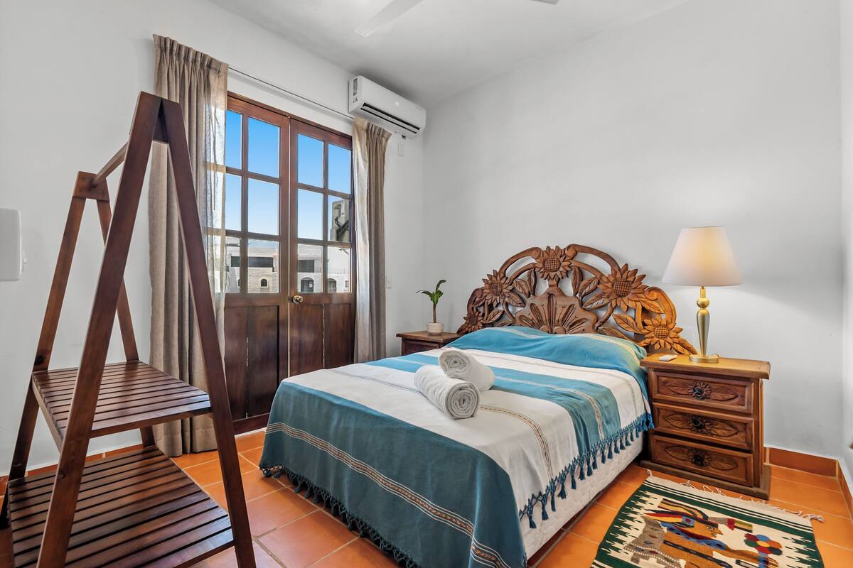 A bedroom with a wooden bed, a wooden ladder, a nightstand with a lamp, a potted plant, and a window with curtains.