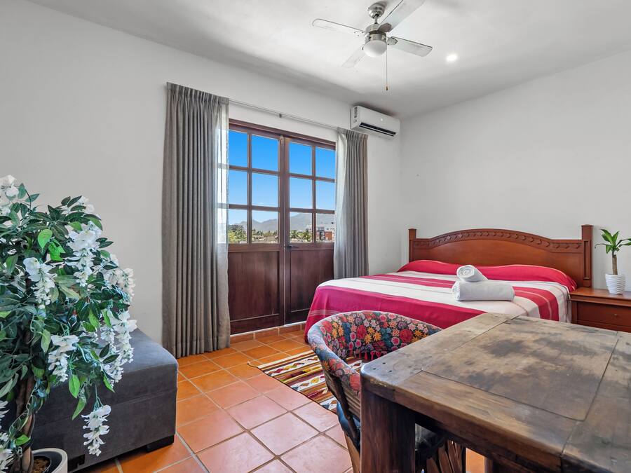 A bedroom with a bed, a desk, a chair, a potted plant, and a window with curtains.