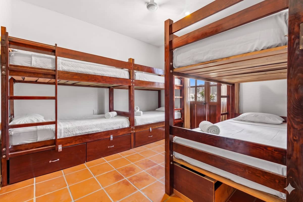 Una habitación con cuatro literas de madera, cada una con colchón y almohadas, y piso de baldosas.