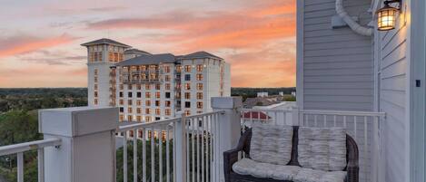 A balcony with a wicker chair and a table, overlooking a multi-story building with balconies, under a sunset sky.