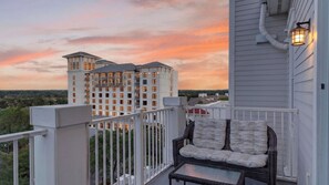 A balcony with a wicker chair and a table, overlooking a multi-story building with balconies, under a sunset sky. - The Grand Grove by Avantstay Beach Access + Pool + Golf + Tennis Walk to Baytowne Wharf (Miramar Beach)