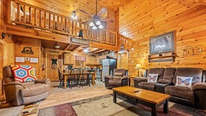 Et interiør af en hytte med et spiseområde, to sofaer, et sofabord og et køkken.