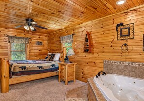 Ein Blockhaus-Interieur mit Bett, Schreibtisch, Lampe, einem Fenster mit Jalousien, Deckenventilator und einer Badewanne.