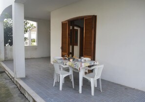 Eine überdachte Terrasse mit einem Tisch für vier Personen, weißen Plastikstühlen und einer Holztür mit Fensterläden.