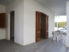 Ein Zimmer mit Fliesenboden, einer weißen Wand und einer Holztür. Ein Tisch mit Stühlen und ein Auto sind draußen geparkt.