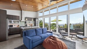 A modern kitchen with a central island, a blue sofa, and a large sliding glass door leading to an outdoor patio with seating. - Trinity Vista by Avantstay Private Dual-zone Spa + Fire Pit + Outdoor Dining + Views Sonoma Wine Country (Glen Ellen)
