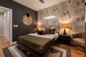 A bedroom with a bed, two bedside tables with lamps, a chair, a decorative wall piece, and a door. - Brick Haven / Hot Tub, Cold Plunge & Sauna Vibes (Nashville)