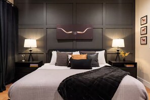 Una habitación de hotel con una cama grande, dos mesitas de noche con lámparas, un cabecero oscuro y una obra de arte colgada en la pared.