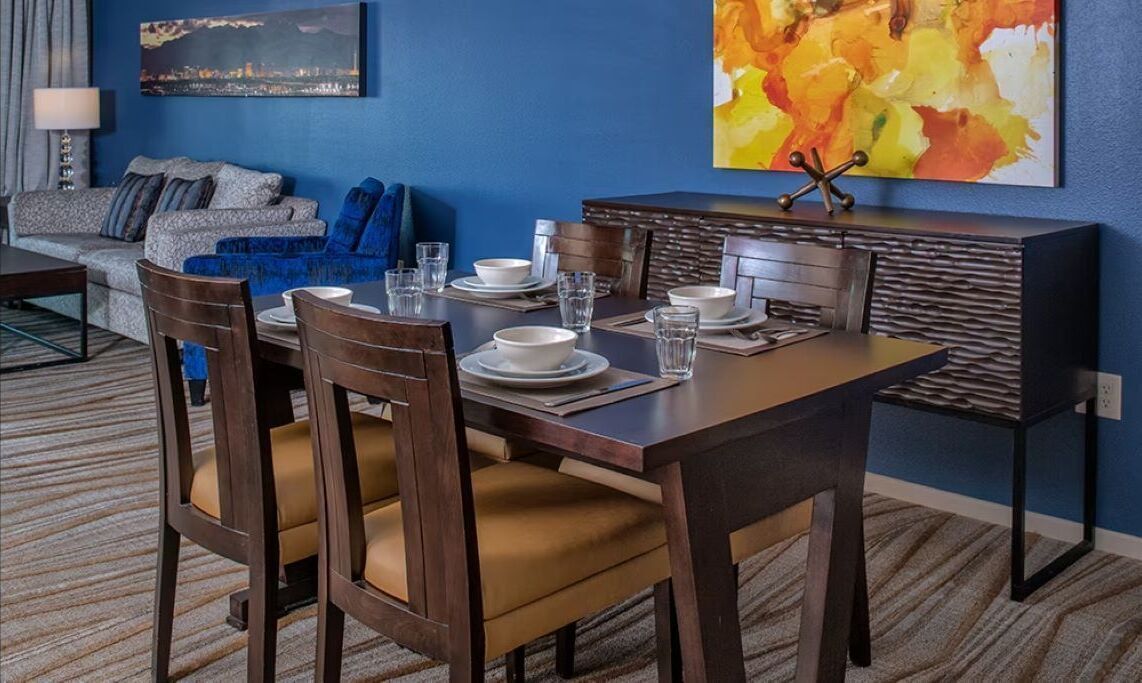 A dining area with a rectangular table set for four, surrounded by wooden chairs with cushioned seats.