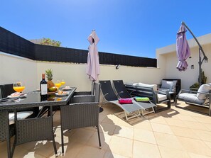 Eine Terrasse im Freien mit einem für das Frühstück gedeckten Tisch, inklusive einer Weinflasche, Obst und Gläsern. Bequeme Liegestühle und Sonnenschirme sind ebenfalls vorhanden.
