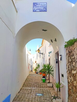 Eine enge Kopfsteinpflasterstraße mit weißen Gebäuden, einem Bogen und einem Schild mit der Aufschrift 'Old Fisherman's Corner'.