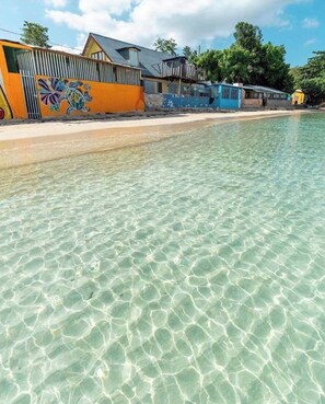 Una platja amb aigua transparent i poc profunda, un mural colorit a un edifici, i algunes construccions al fons.