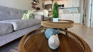 Ein Wohnzimmer mit grauem Sofa, einem Holztisch mit dekorativen Fischen, einem weißen Sessel und einer Küche im Hintergrund.