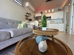 Un saló amb un sofà gris, una taula de centre de fusta amb peixos decoratius, una cadira blanca i una cuina al fons.