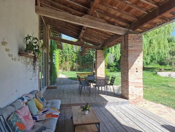 A covered patio with a wooden table and chairs, a sofa, and a brick wall.
