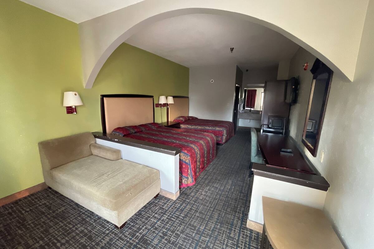 A hotel room with a sofa, two beds with red bedding, a desk, a mirror, and a television.