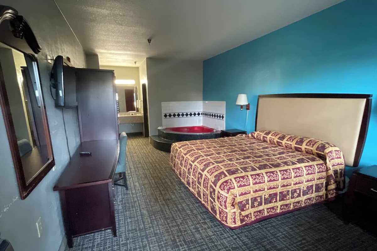 A hotel room with a bed, a desk, a chair, a mirror, a bathroom with a bathtub, and a wall-mounted lamp.