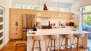 Une cuisine moderne avec des meubles en bois, un grand îlot central doté de tabourets, un éclairage suspendu et une vue sur l’extérieur.