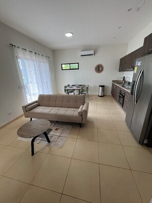 Un saló amb un sofà, una taula de dinar i cadires, una cuina amb nevera, i una finestra amb cortines.