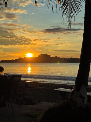 A sunset over a calm sea with a silhouette of a cityscape in the distance, a beach chair, and a palm tree. - Charming 2-bedroom 2-bath condo with AC in Potrero 5-7 minutes from the beach (Potrero)