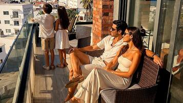 A balcony with a glass railing, wicker furniture, and a view of buildings and hills.