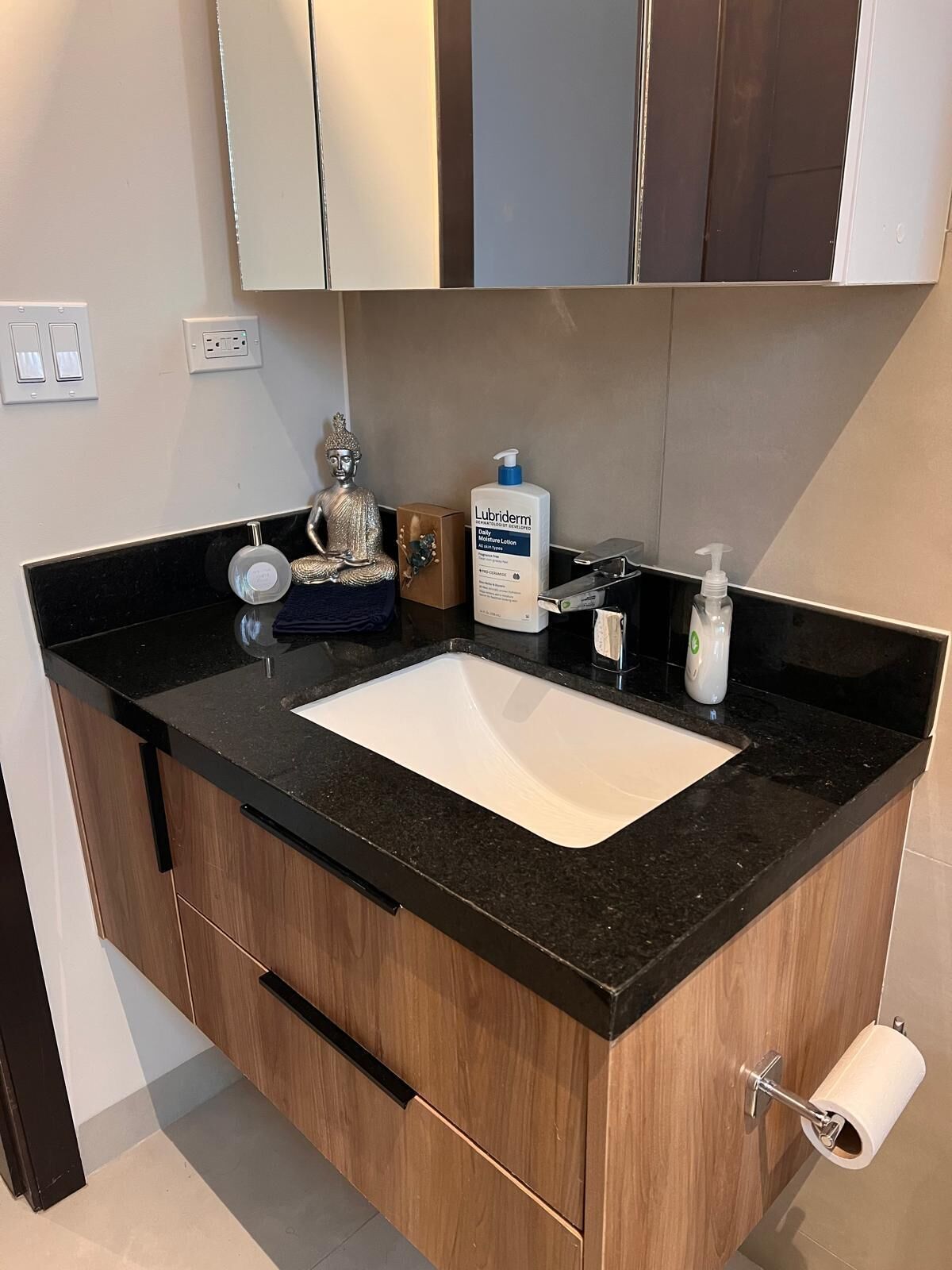 A bathroom with a black countertop, a white sink, a mirror, a Buddha statue, a bottle of Lubriderm lotion, and a toilet paper holder.