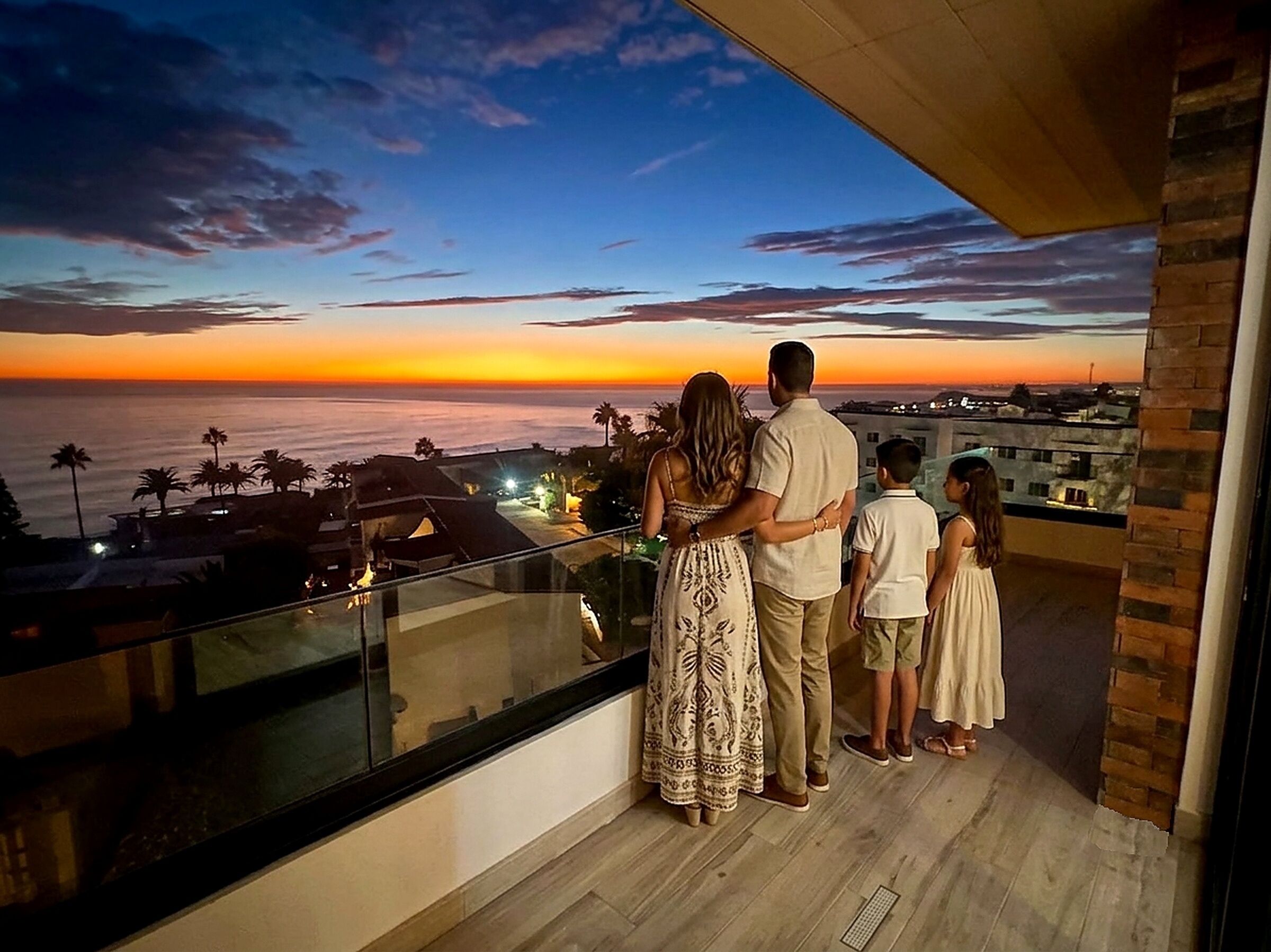 A balcony with a view of the sunset over the ocean, a family standing together, and a modern building in the background.