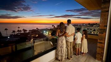 A balcony with a view of the sunset over the ocean, a family standing together, and a modern building in the background.