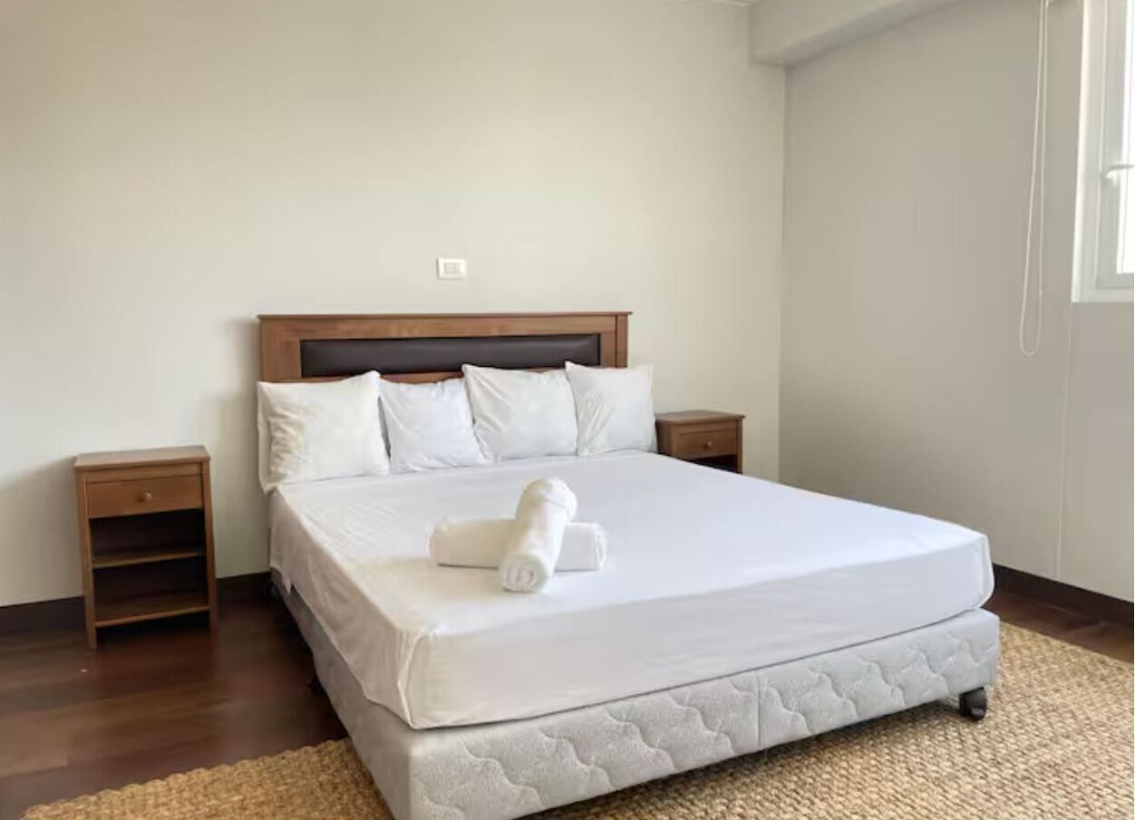 Een net opgemaakt bed met een opgerolde handdoek, een houten hoofdbord en twee nachtkastjes.